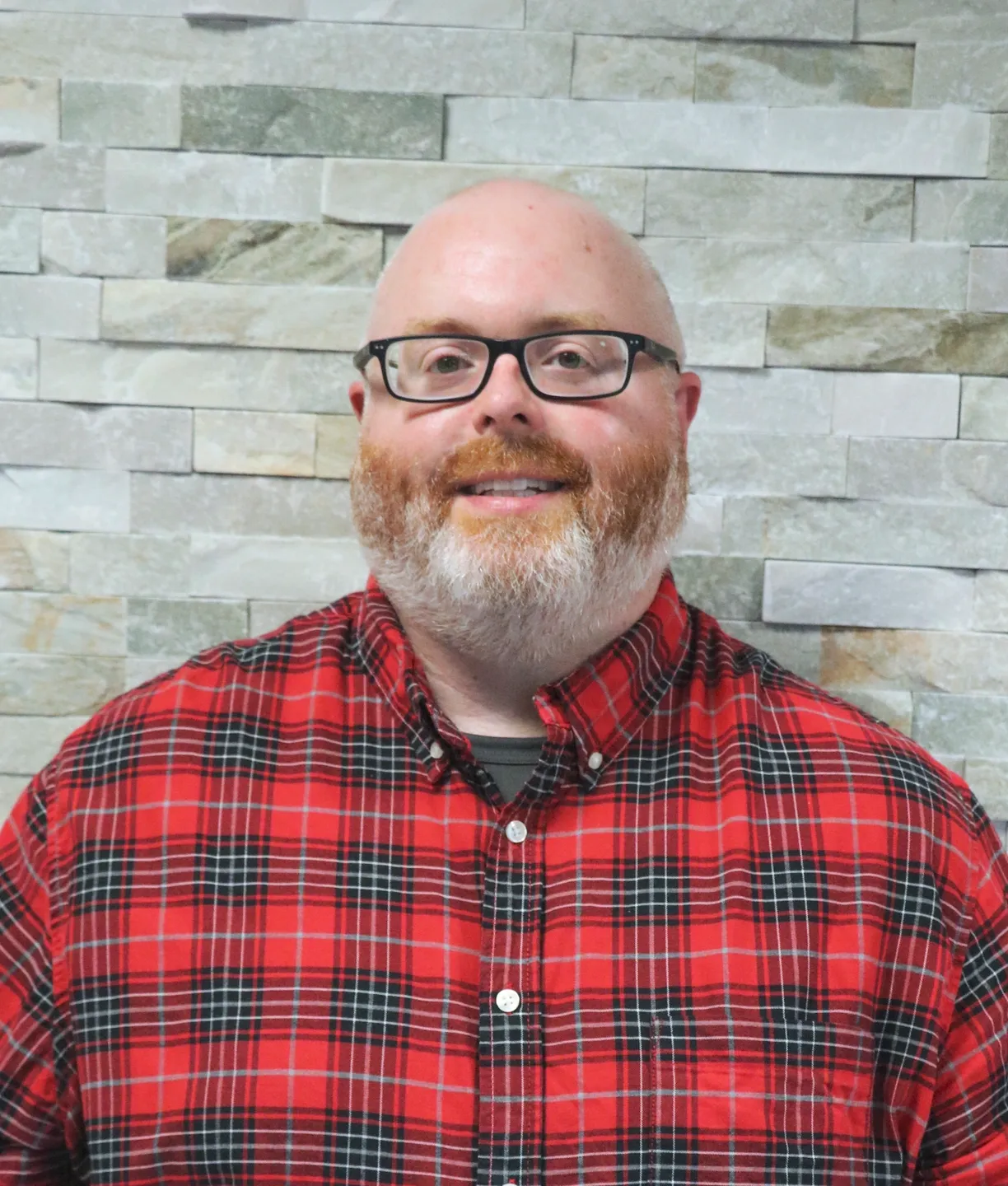A bald man in a red plaid shirt standing in front of a stone wall.