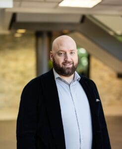 A man with a beard and jacket standing in front of a building.